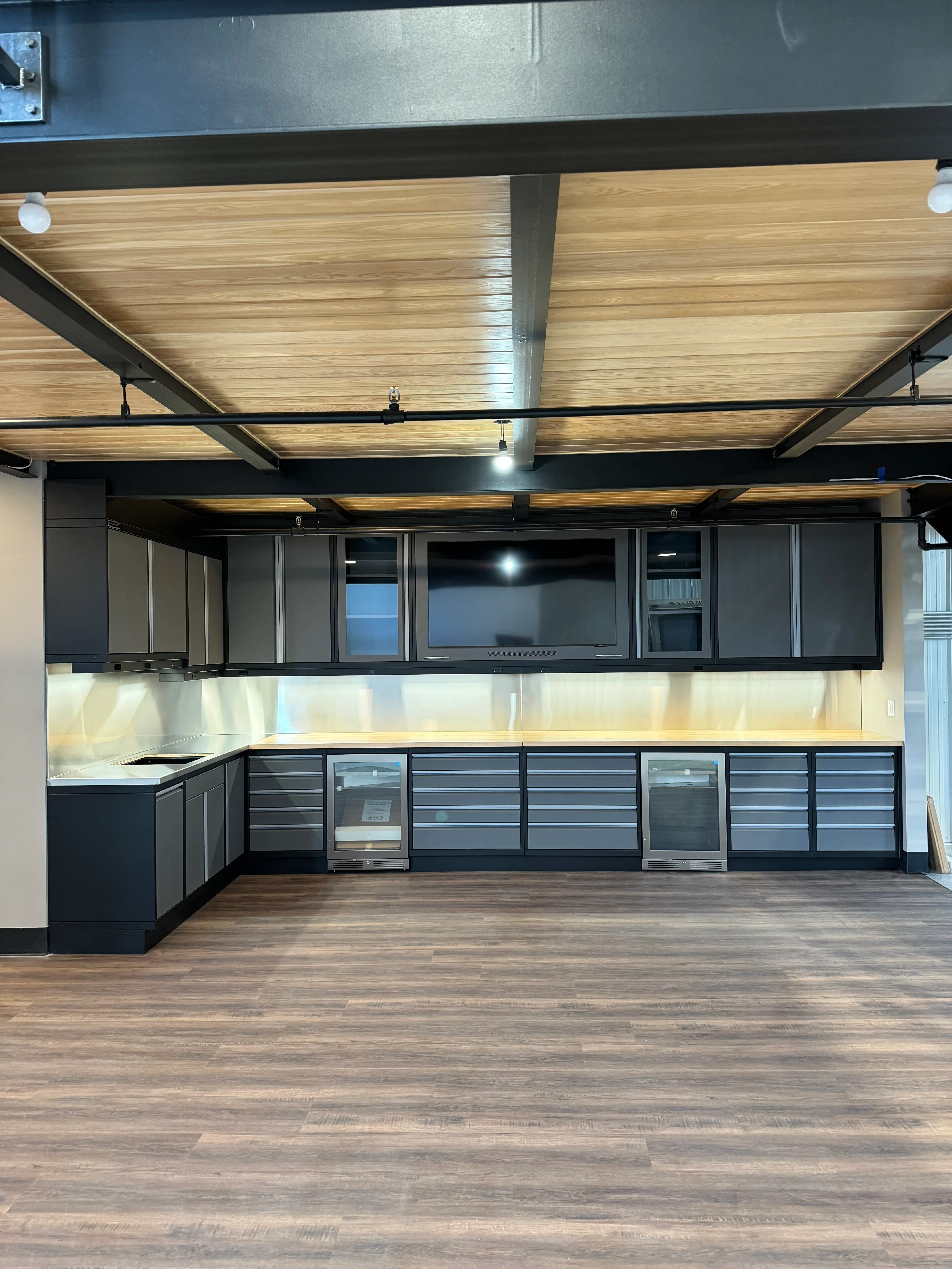 Modern custom cabinet system by Baldhead Cabinets featuring matte gray and black powder-coated finish, under-cabinet lighting, butcher block countertops, dual beverage refrigerators, wall-mounted TV, and wood plank flooring in a contemporary garage or showroom space.