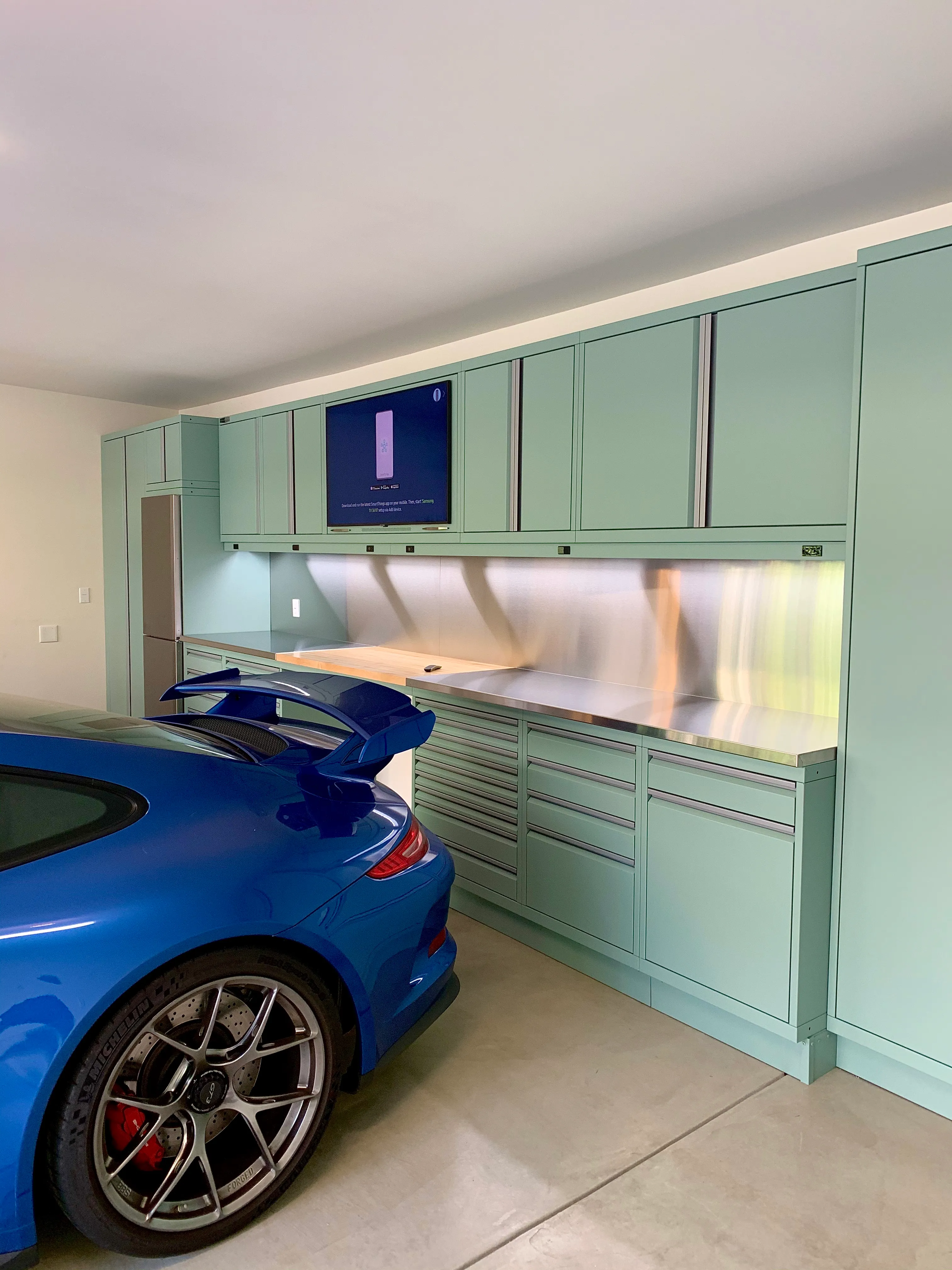 Sleek mint green Baldhead Cabinets with stainless steel countertops and integrated TV, paired with a luxury blue Porsche for a high-end garage setup combining performance, style, and precision craftsmanship.