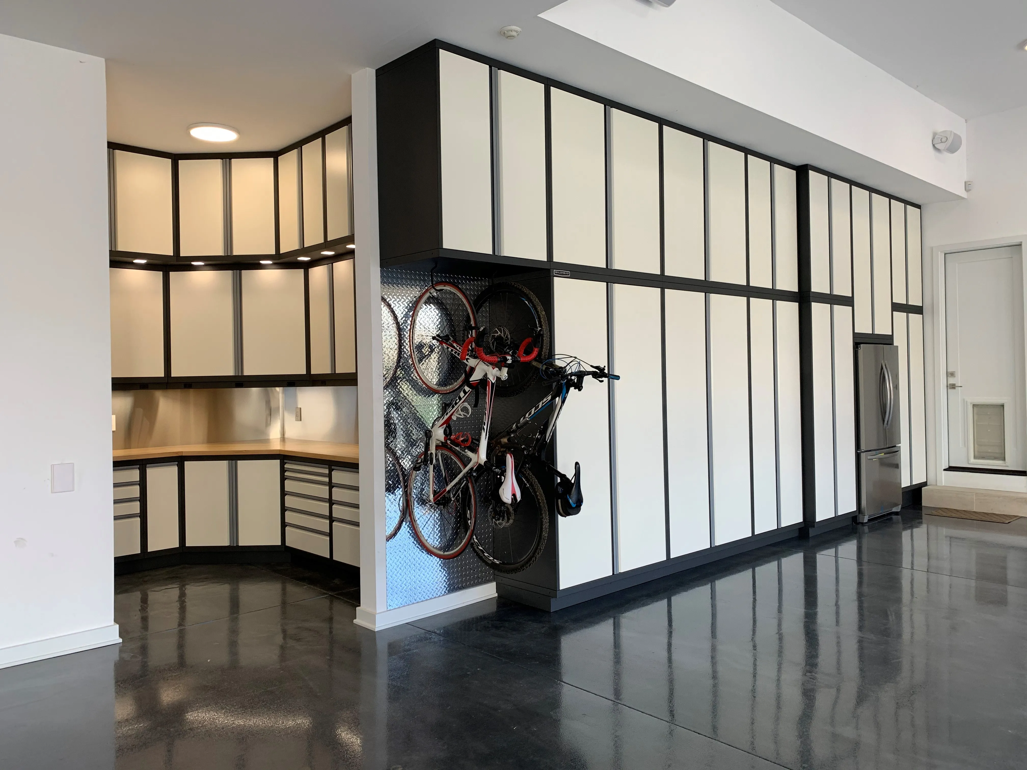 Premium garage storage system by Baldhead Cabinets featuring two-tone cream and black powder-coated cabinetry, corner workstation with under-cabinet lighting, integrated stainless steel refrigerator, vertical bike rack, and high-gloss black epoxy flooring in a luxury home garage.